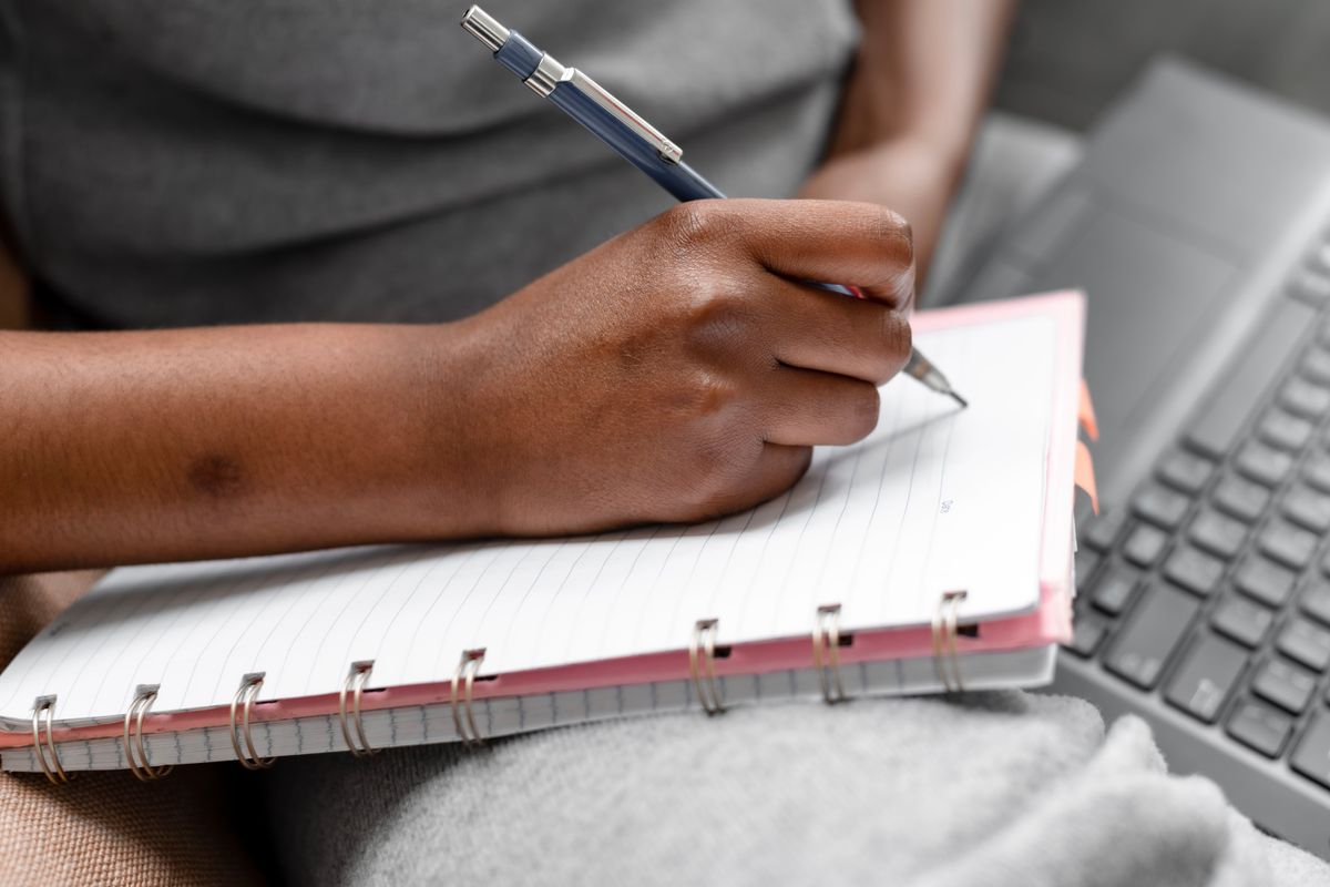 A person writing in a notebook next to a laptop